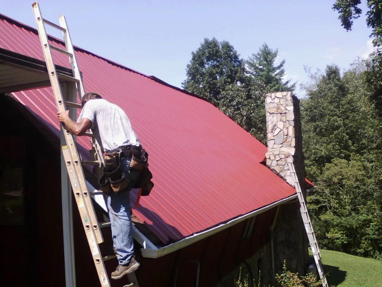 Roofer on a ladder beside a red metal roof and stone chimney — Soffit & Fascia Repair by Roofing Brothers in Oakpark